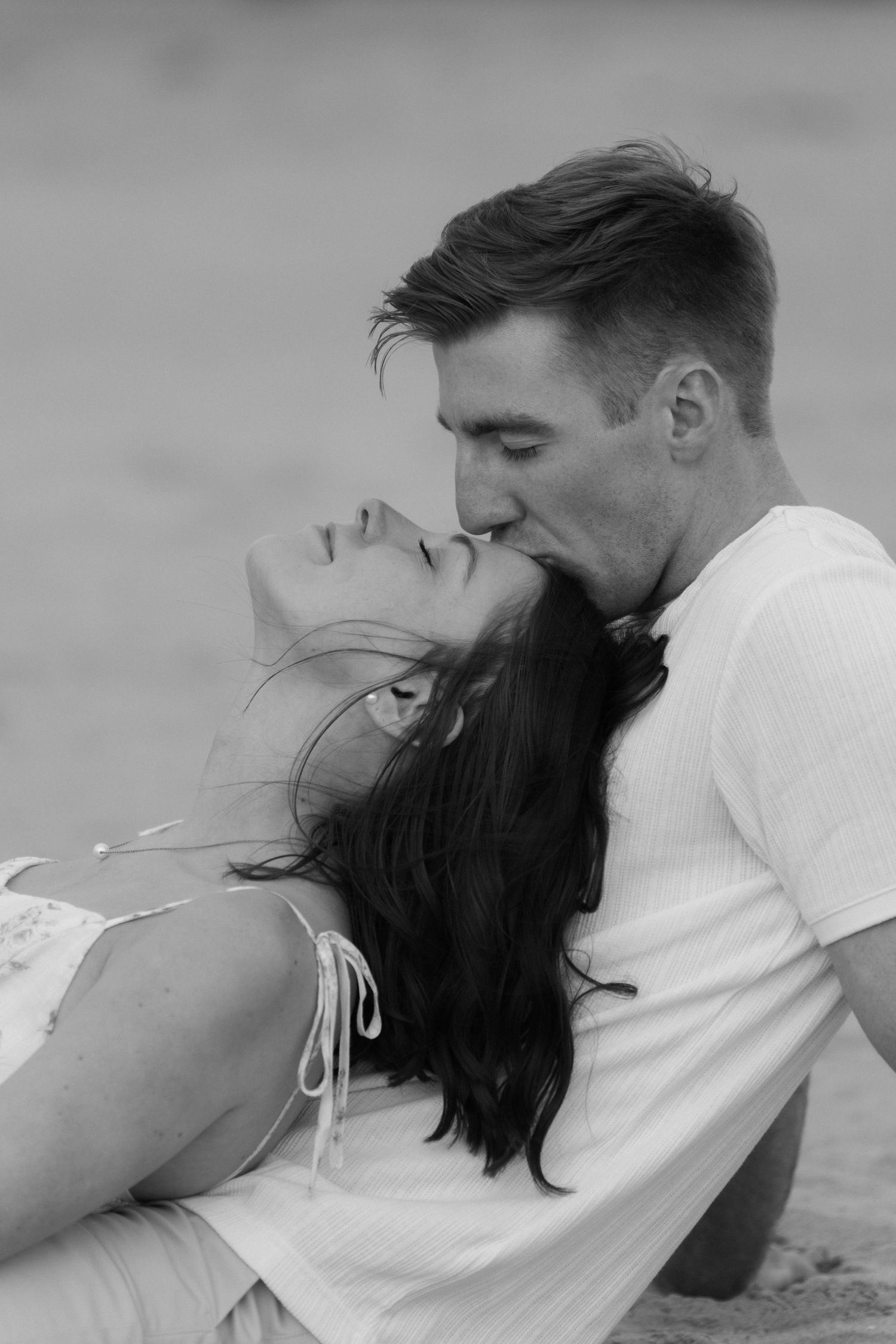 Black and white photo of man tenderly kissing woman on forehead while sitting together on a sandy beach.
