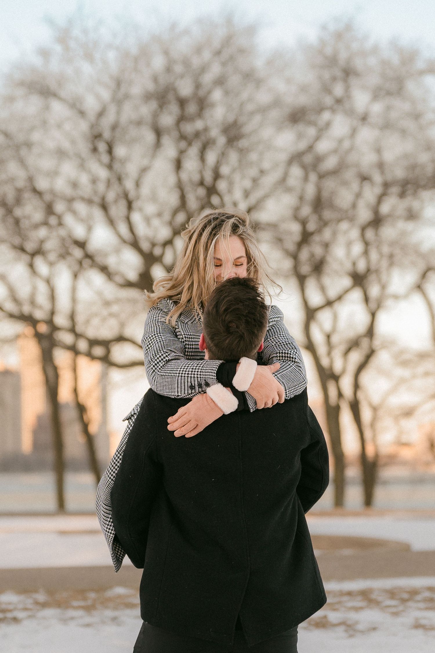 A man lifts a laughing woman in his arms among bare winter trees at sunset.