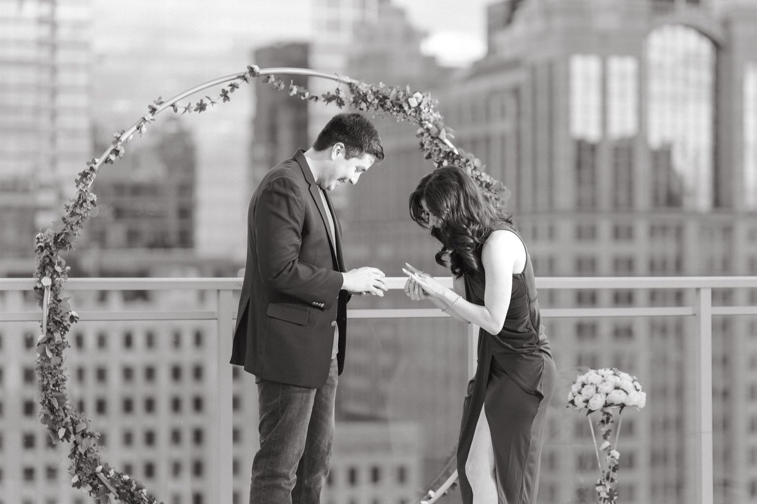 Black and white photo of couple examining engagement ring on urban rooftop with city skyline backdrop.