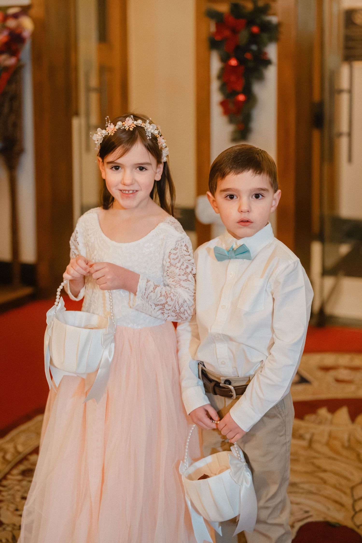 Adorable flower girl in pink dress and ring bearer in bow tie hold white baskets at church wedding ceremony.