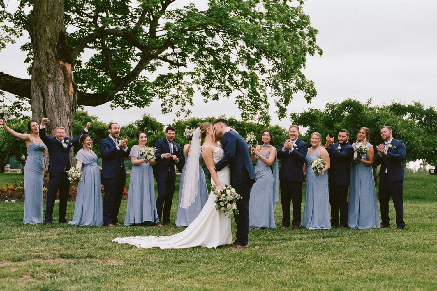 Wedding party poses outdoors under large tree, bride and groom kissing while bridesmaids in blue gowns and groomsmen in navy suits cheer.