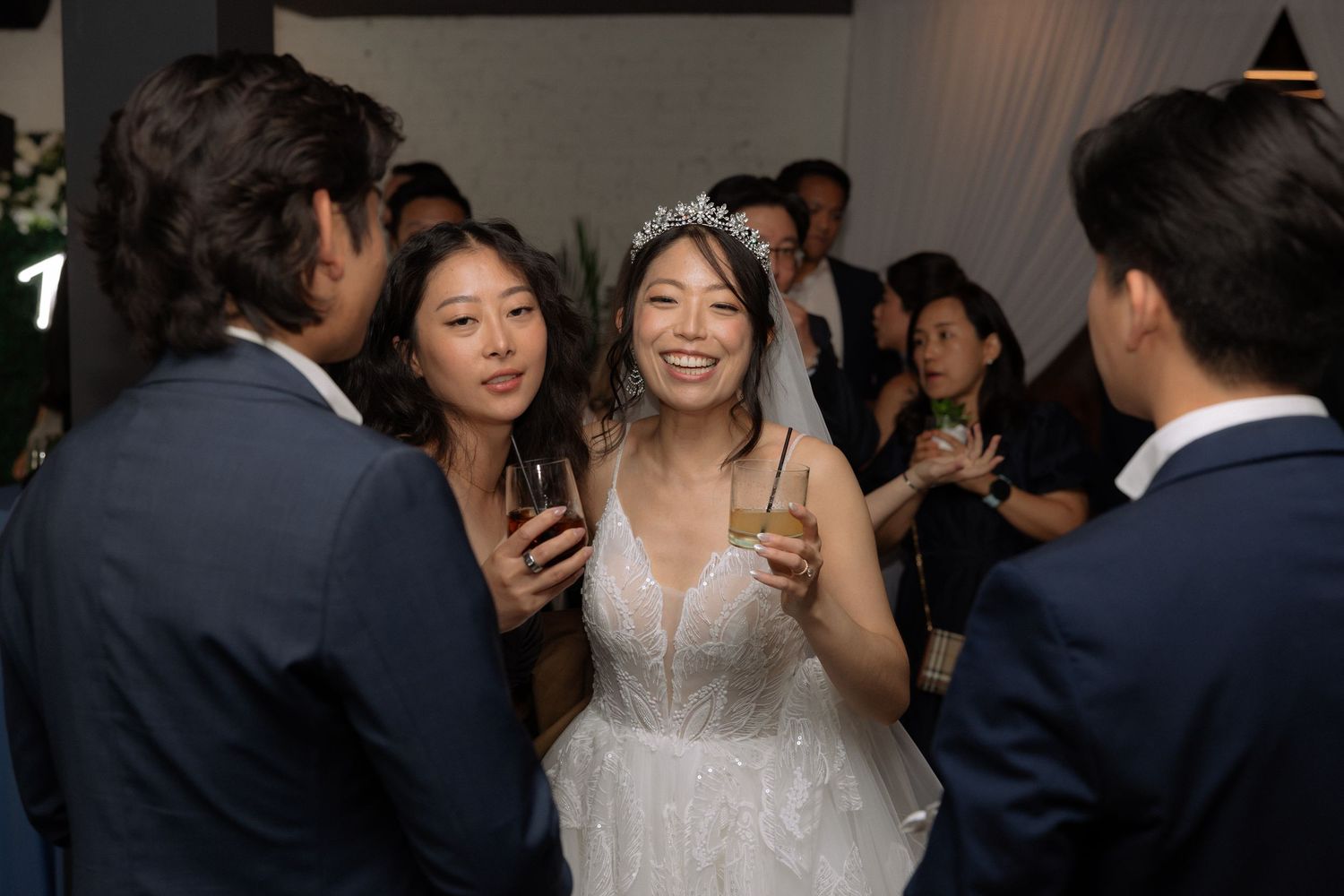 Smiling bride in white gown and tiara holds champagne glass while chatting with guests at elegant wedding reception.