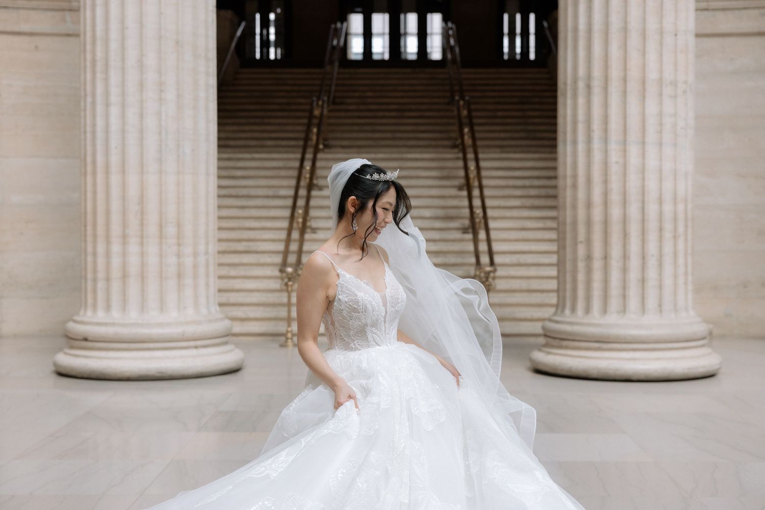 Bride in stunning ballgown wedding dress with long veil poses between grand columns in ornate venue.