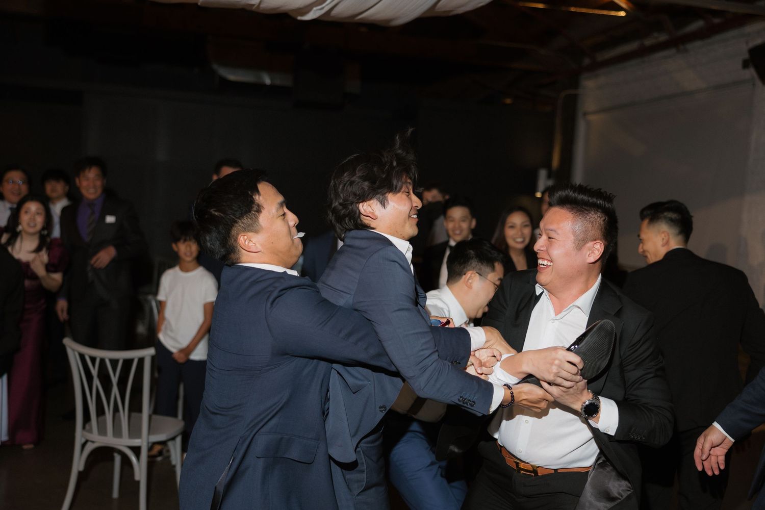 Groomsmen and wedding guests playfully wrestling and laughing together at indoor wedding reception.