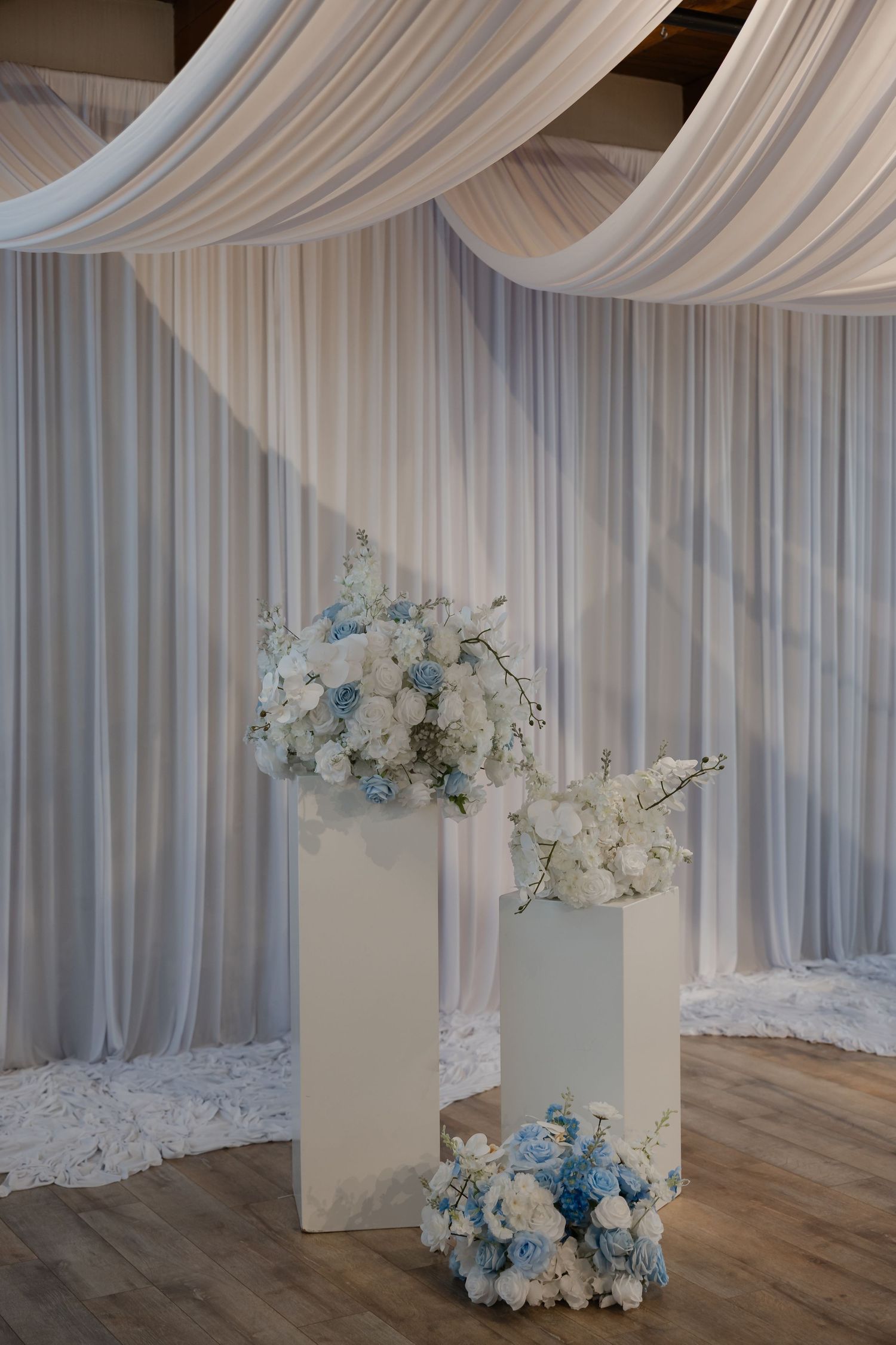 White and blue floral arrangements on tall white pedestals with elegant draped fabric backdrop at a wedding venue.