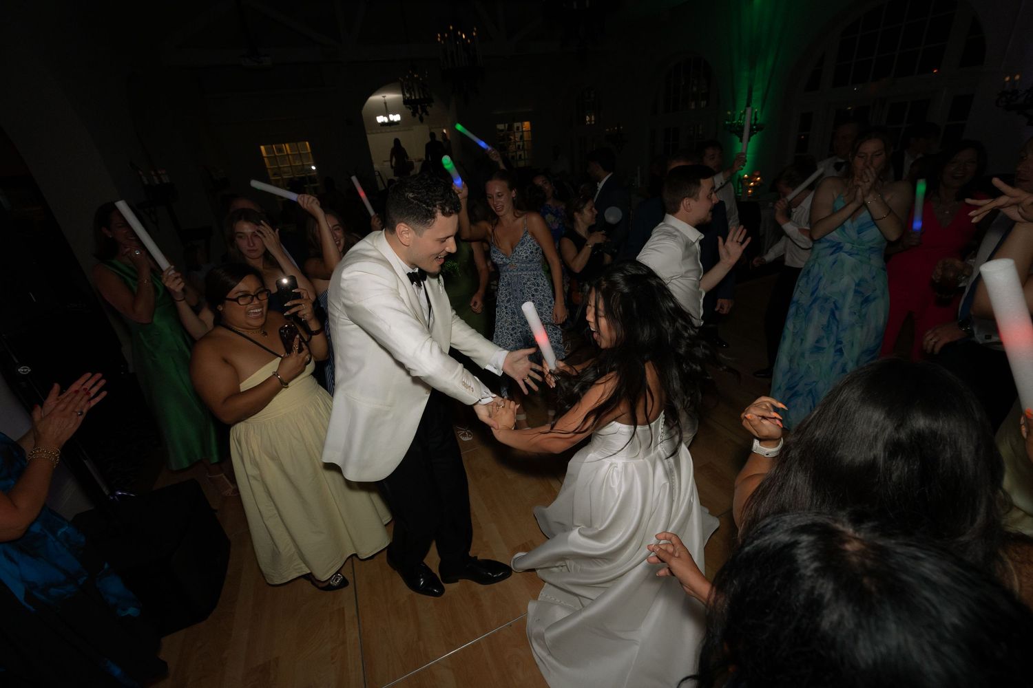 Bride and groom dance energetically with guests on a crowded wedding reception dance floor with colorful lighting.