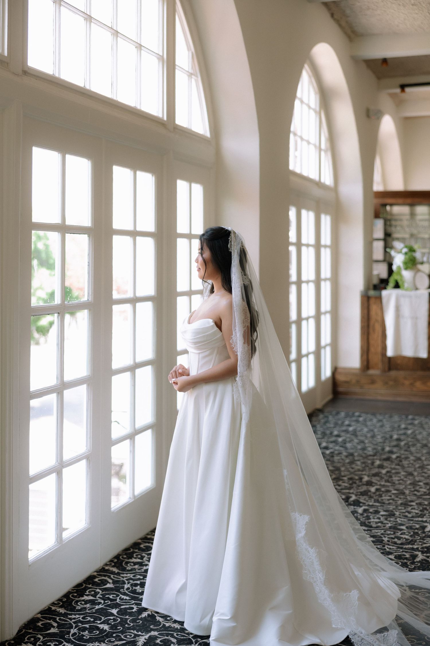 Elegant bride in strapless white gown with cathedral veil standing by arched windows in a bright venue.