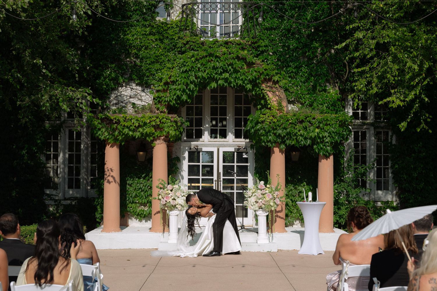Couple sharing first kiss at outdoor wedding ceremony with ivy-covered building, white floral arrangements, and seated guests.