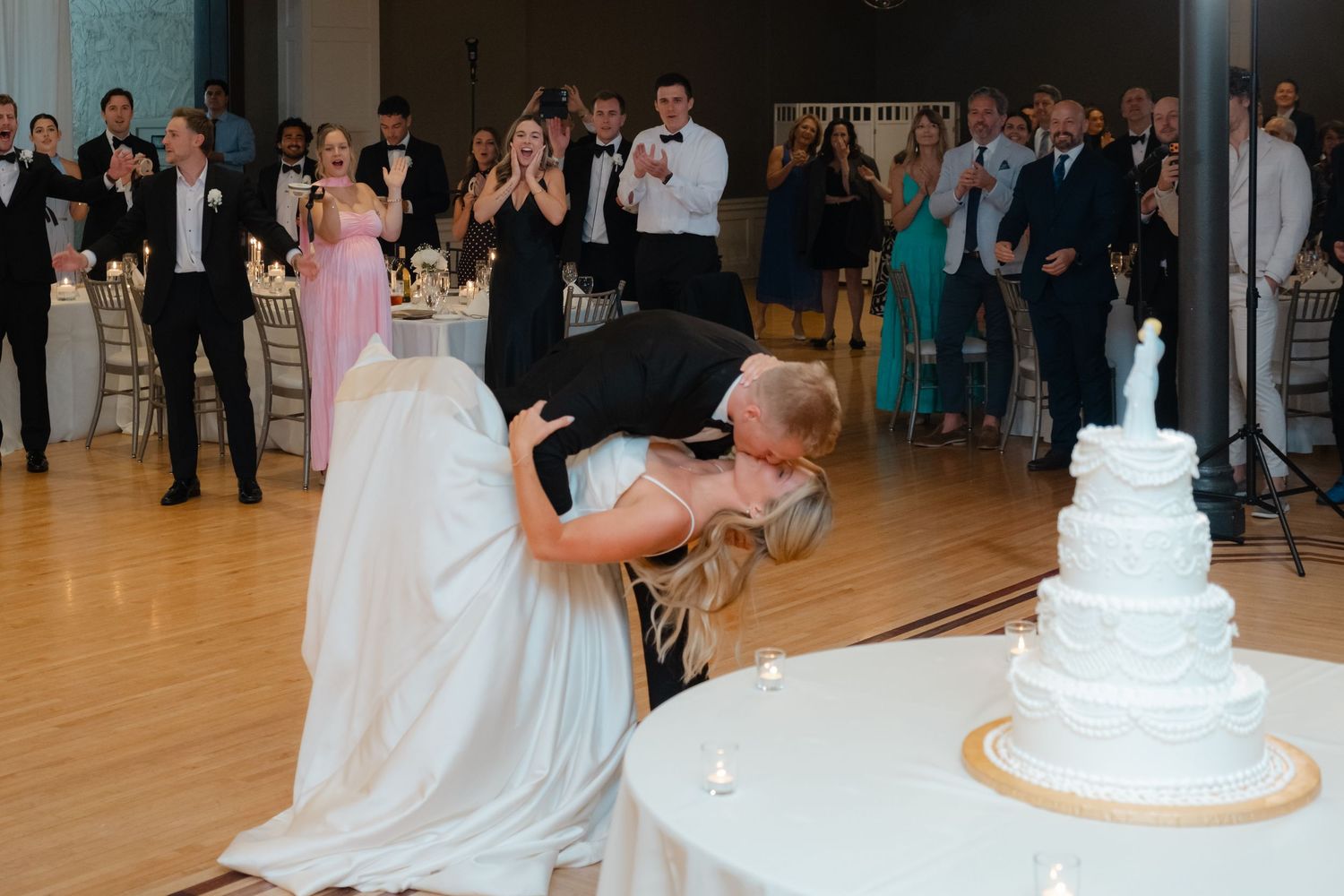 Groom dips bride in dramatic kiss near wedding cake while guests cheer at elegant reception celebration.