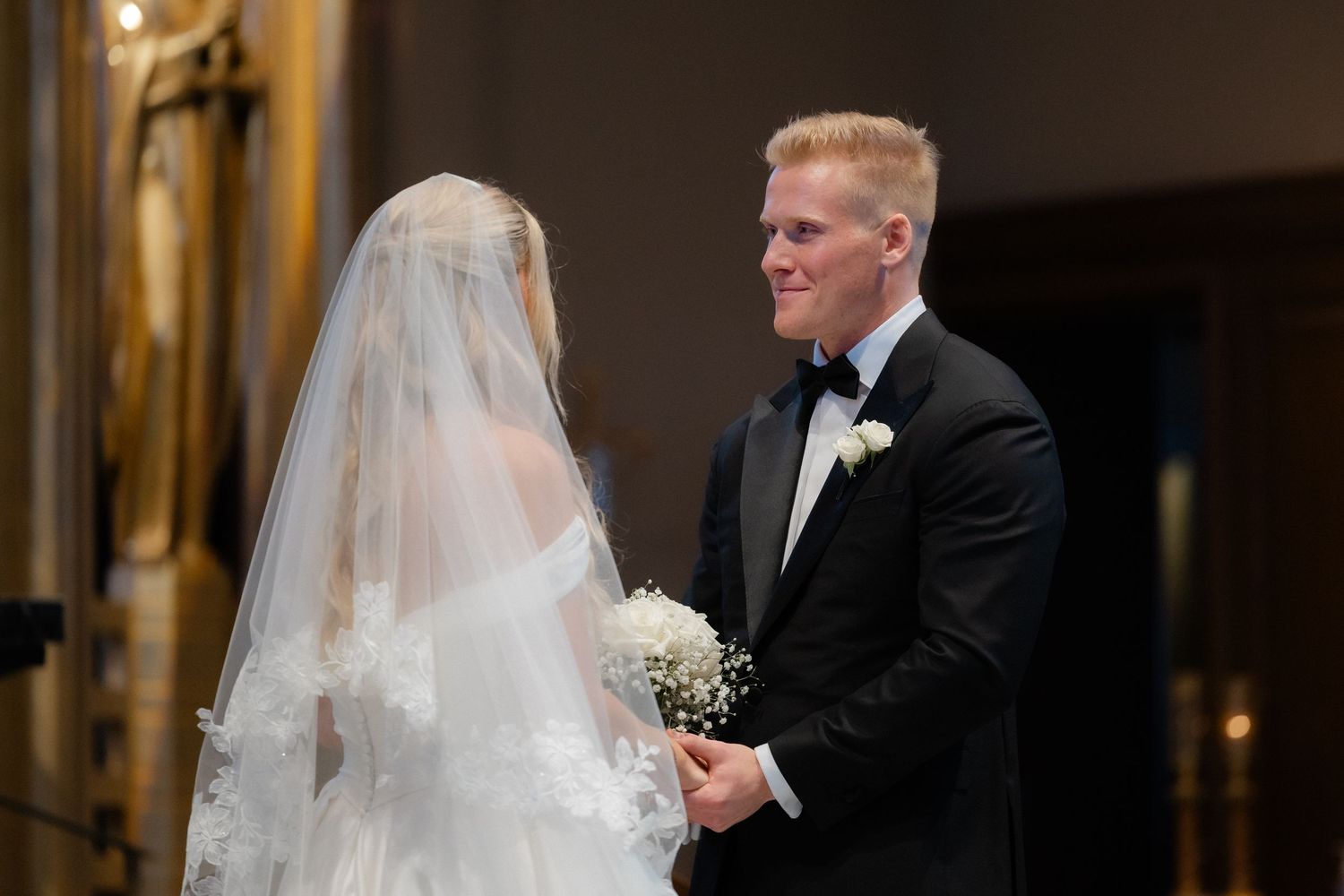 Groom in black tuxedo smiling at veiled bride holding a white bouquet during wedding ceremony.