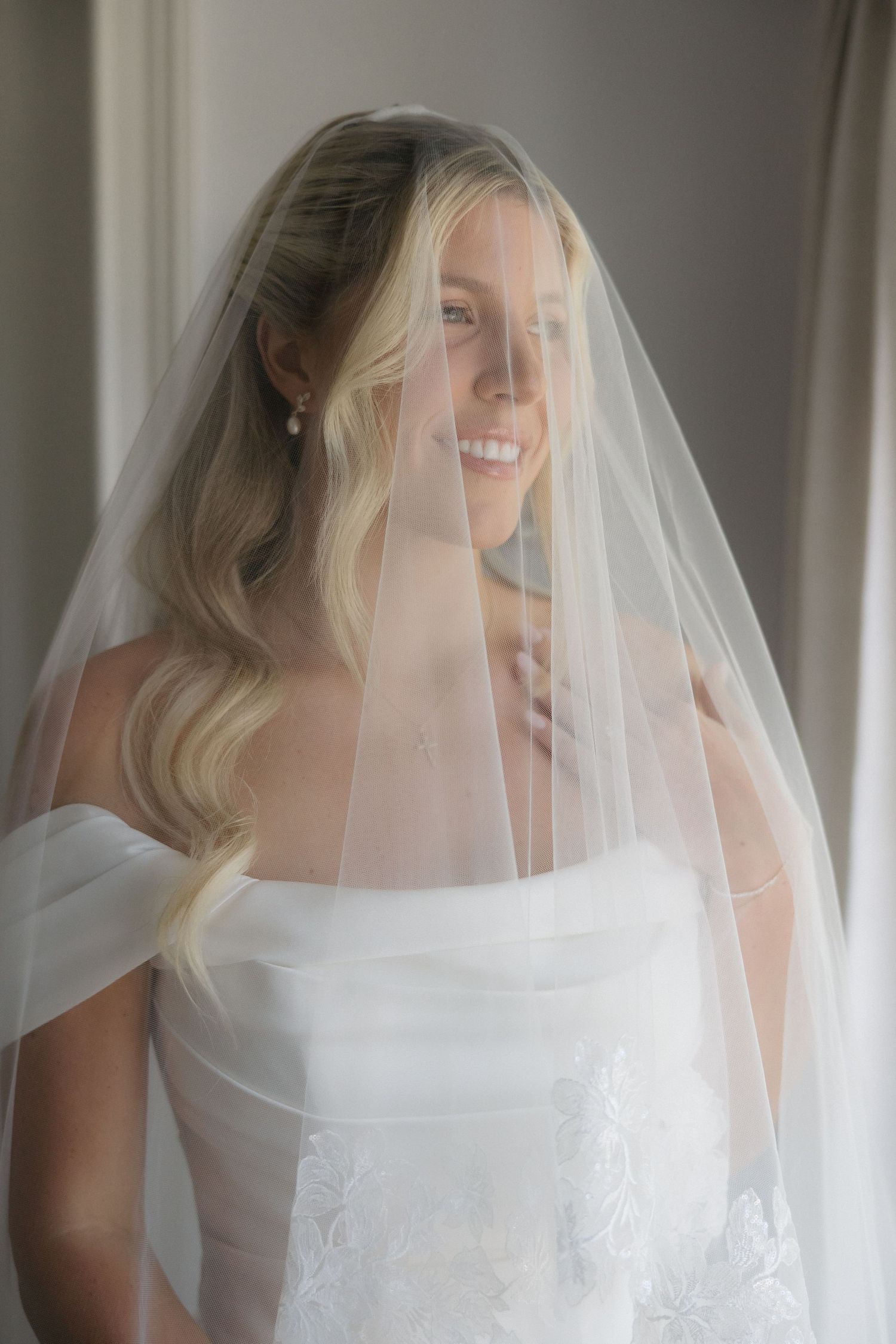 Smiling bride in off-shoulder lace gown and flowing veil with wavy blonde hair and pearl earrings.