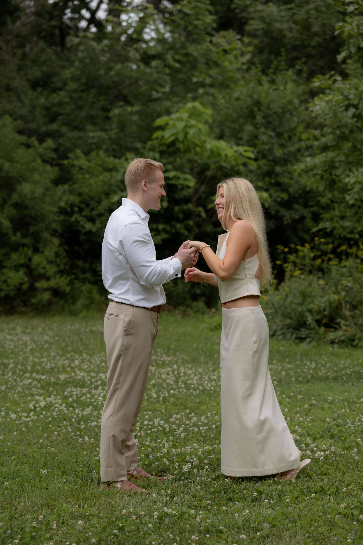 Couple holding hands and smiling while dancing together on a flower-filled lawn surrounded by lush green trees.