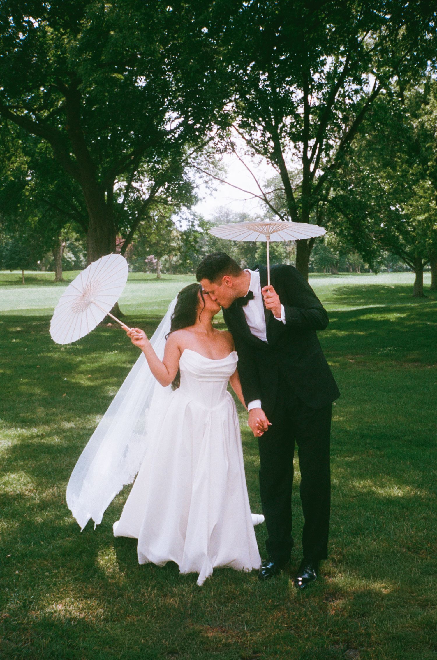Bride and groom kissing while holding white parasols in a lush green park on their wedding day.