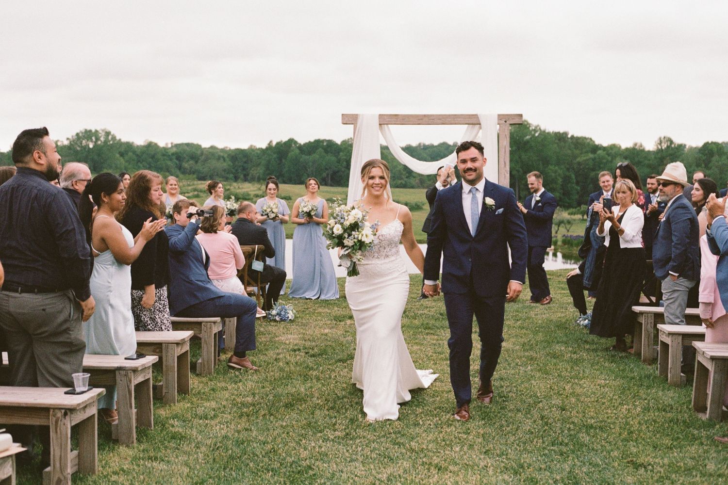 Bride and groom walk down outdoor wedding aisle holding hands, guests seated on wooden benches, rustic arch backdrop.
