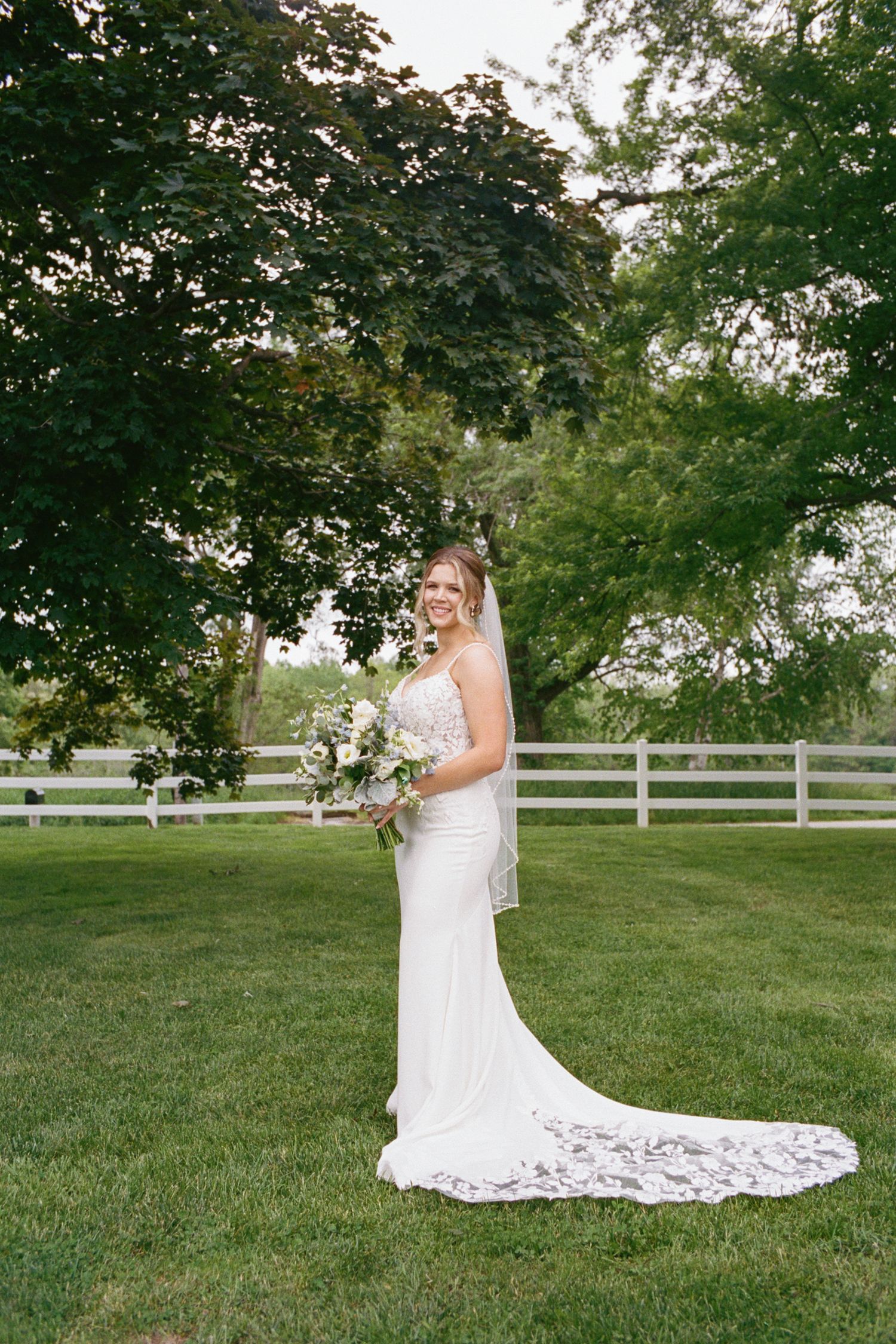 Bride in white lace gown holding bouquet stands on green lawn with white fence and trees in background.