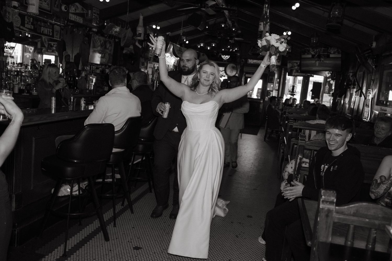 Black and white photo of a joyful bride raising her bouquet in a bar venue during her wedding reception.