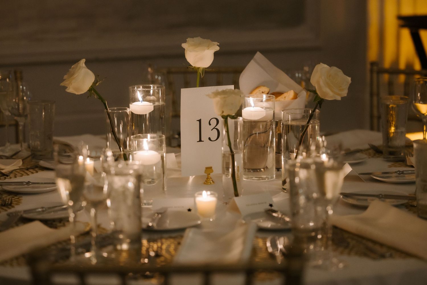 Elegant wedding reception table 13 with white roses, floating candles, and gold accents in a warmly lit ballroom.
