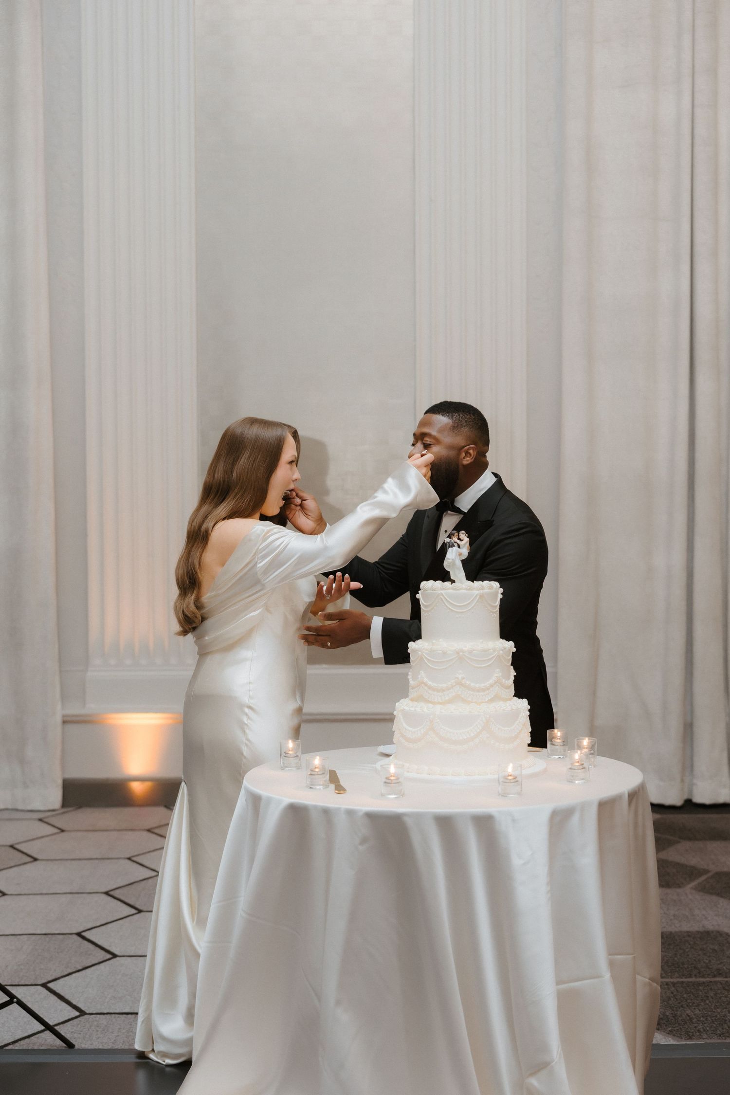 Bride and groom feeding each other wedding cake at elegant reception with white tiered cake and candles.