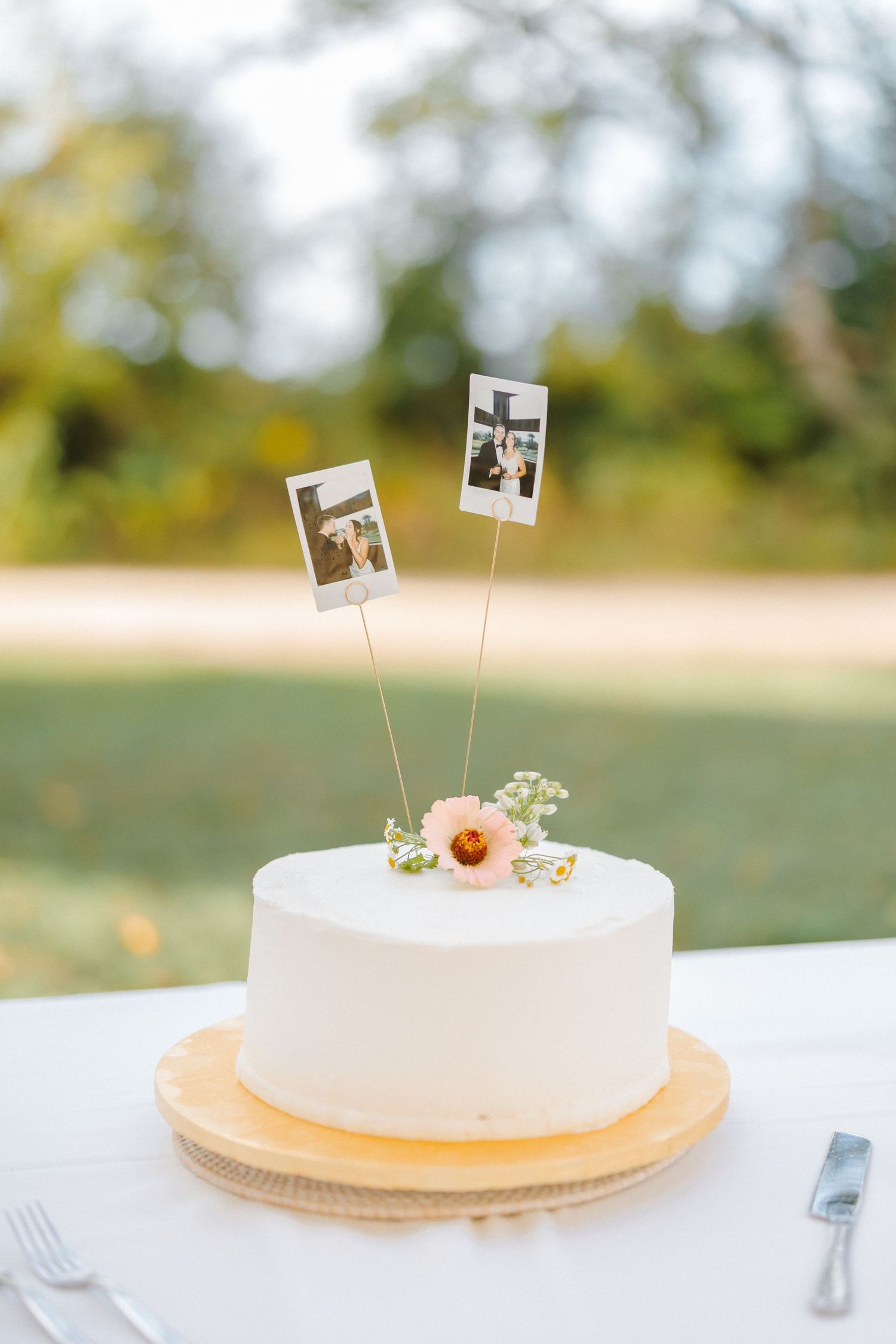 Simple one-tier white wedding cake topped with Polaroid photo cake toppers and a pink flower.