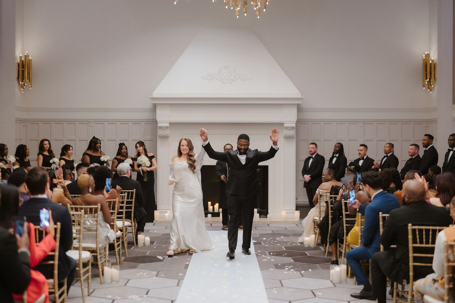 Joyful newlyweds celebrate recessional in elegant white wedding venue with gold chairs and chandelier lighting.