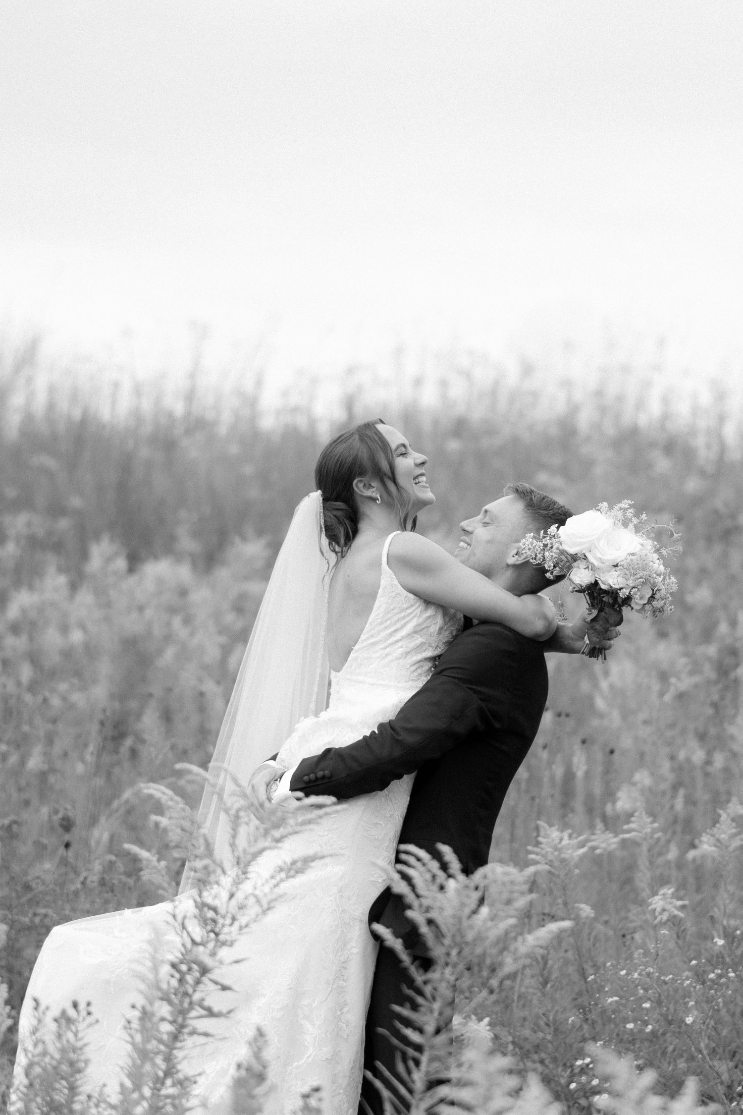 Black and white photo of groom lifting laughing bride in a wildflower field.