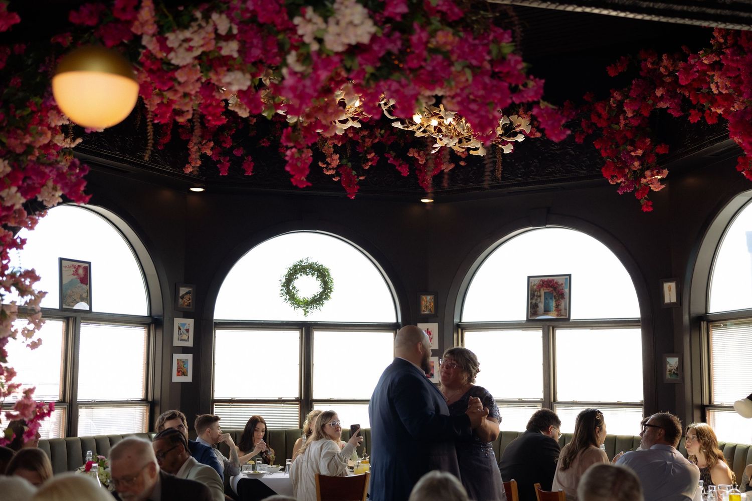 Couple dancing in elegant restaurant with floral ceiling, arched windows, and seated guests at a wedding reception.