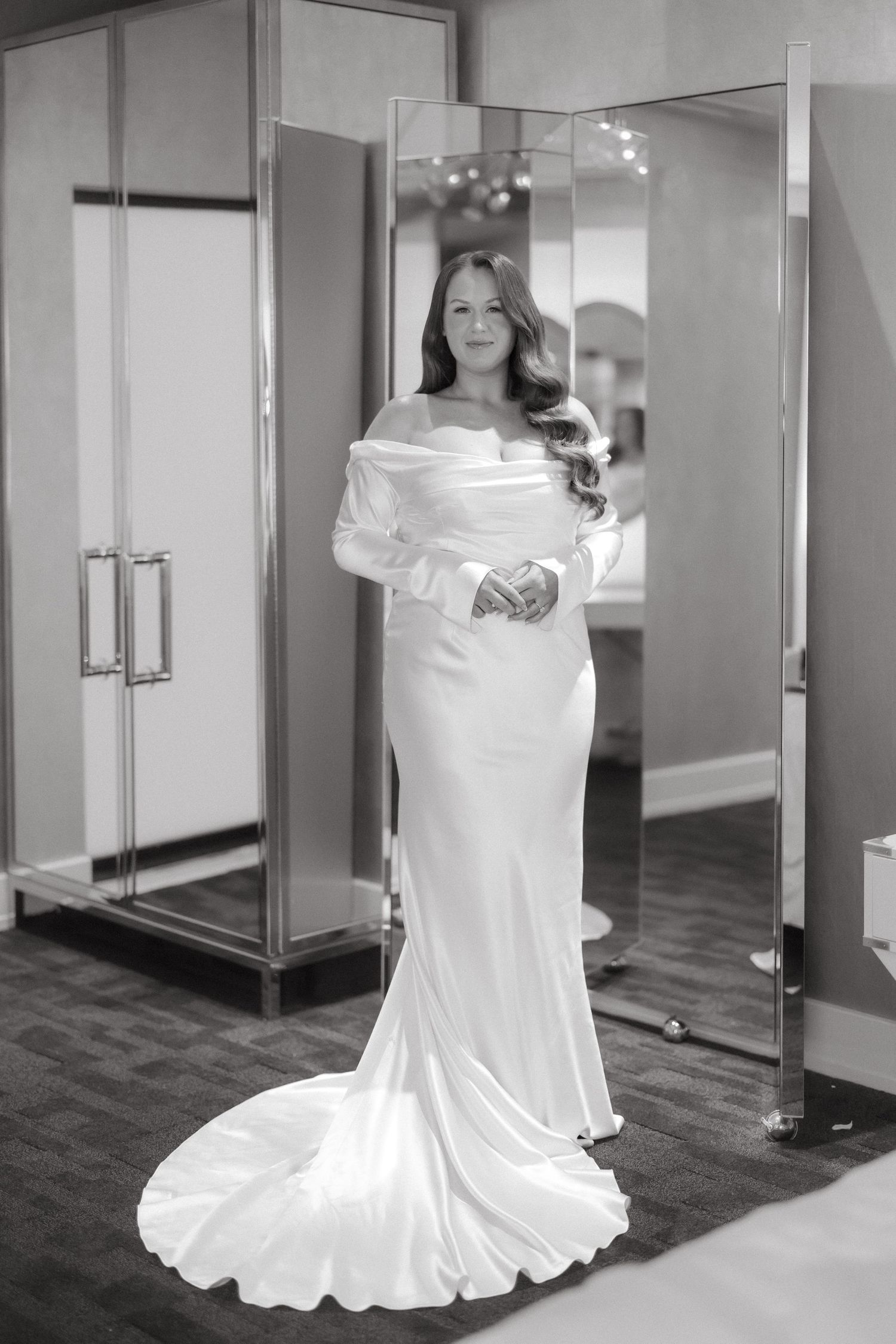 Bride in elegant off-shoulder long-sleeve white satin wedding gown with train, posing in bridal boutique fitting room.