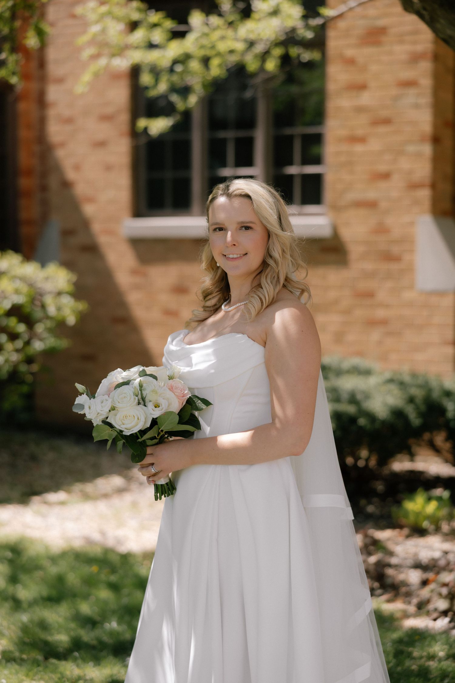 Smiling bride in strapless white gown holding white rose bouquet outdoors near brick church on sunny day.