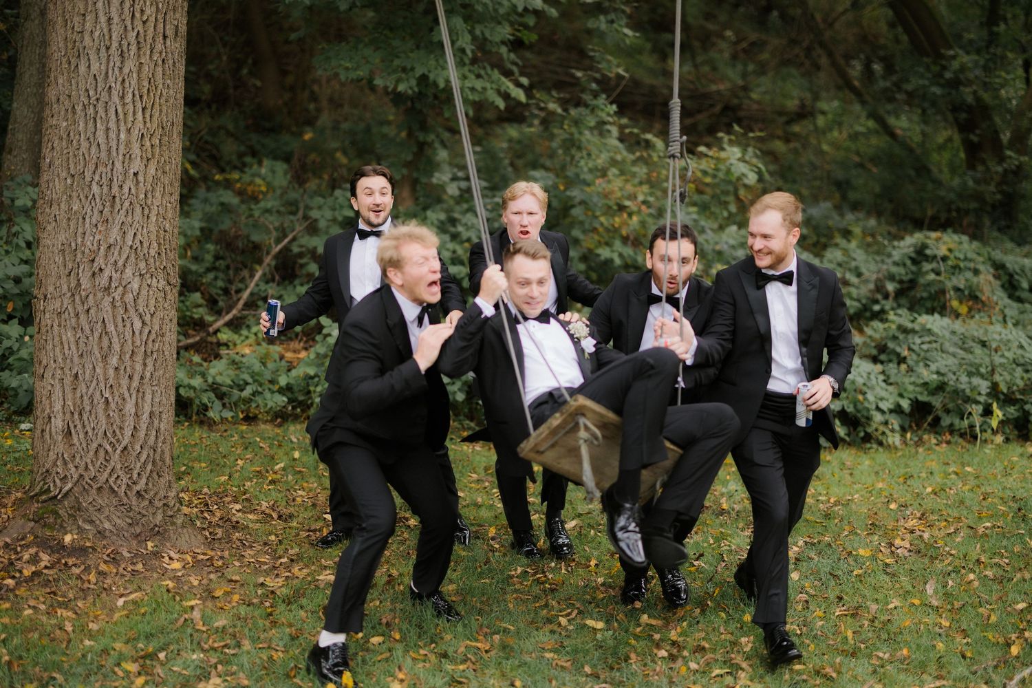 Groomsmen in black tuxedos playfully push groom on wooden swing surrounded by autumn foliage.