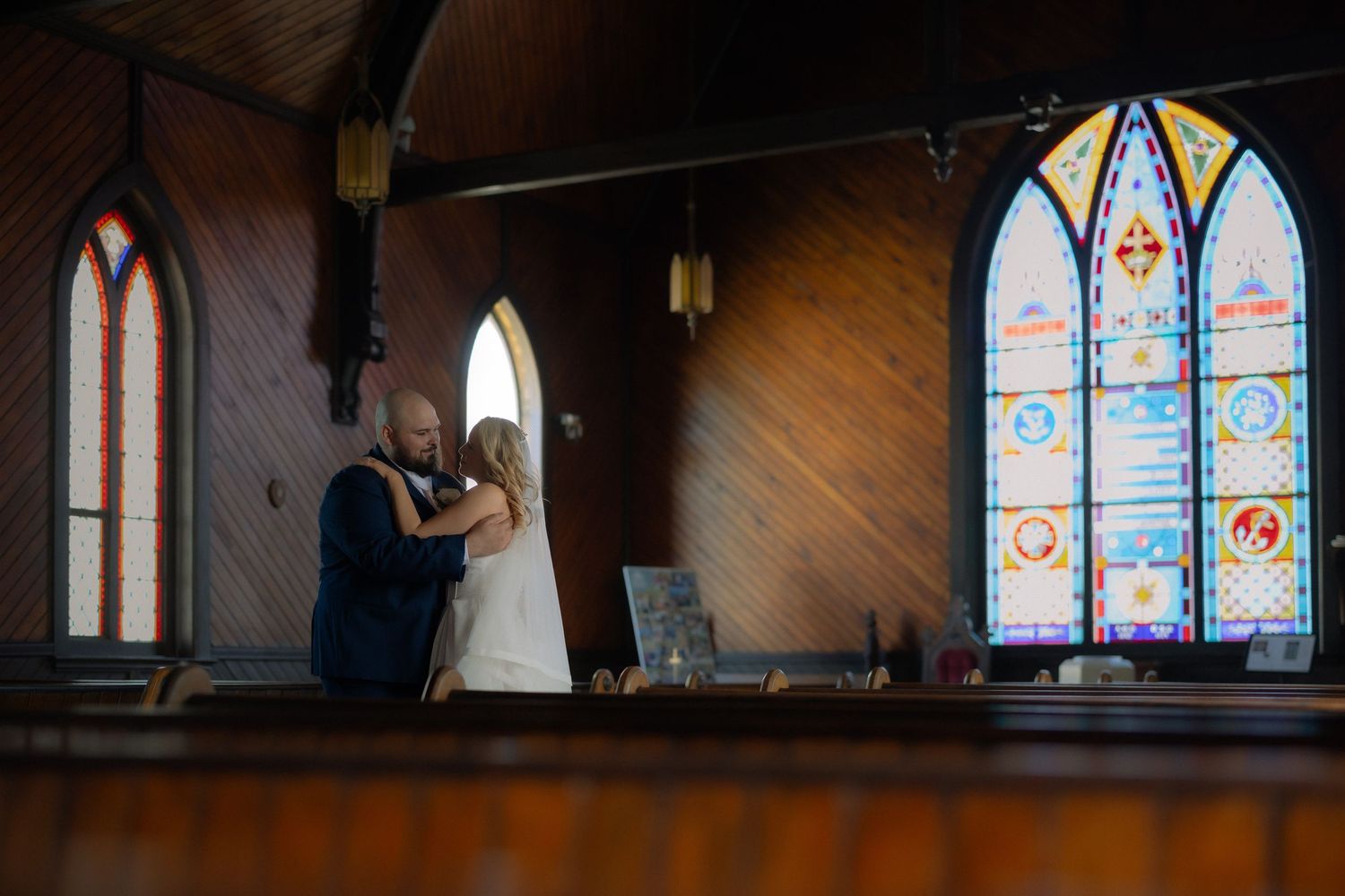 A bride and groom share a romantic embrace inside a wooden church with colorful stained glass windows.