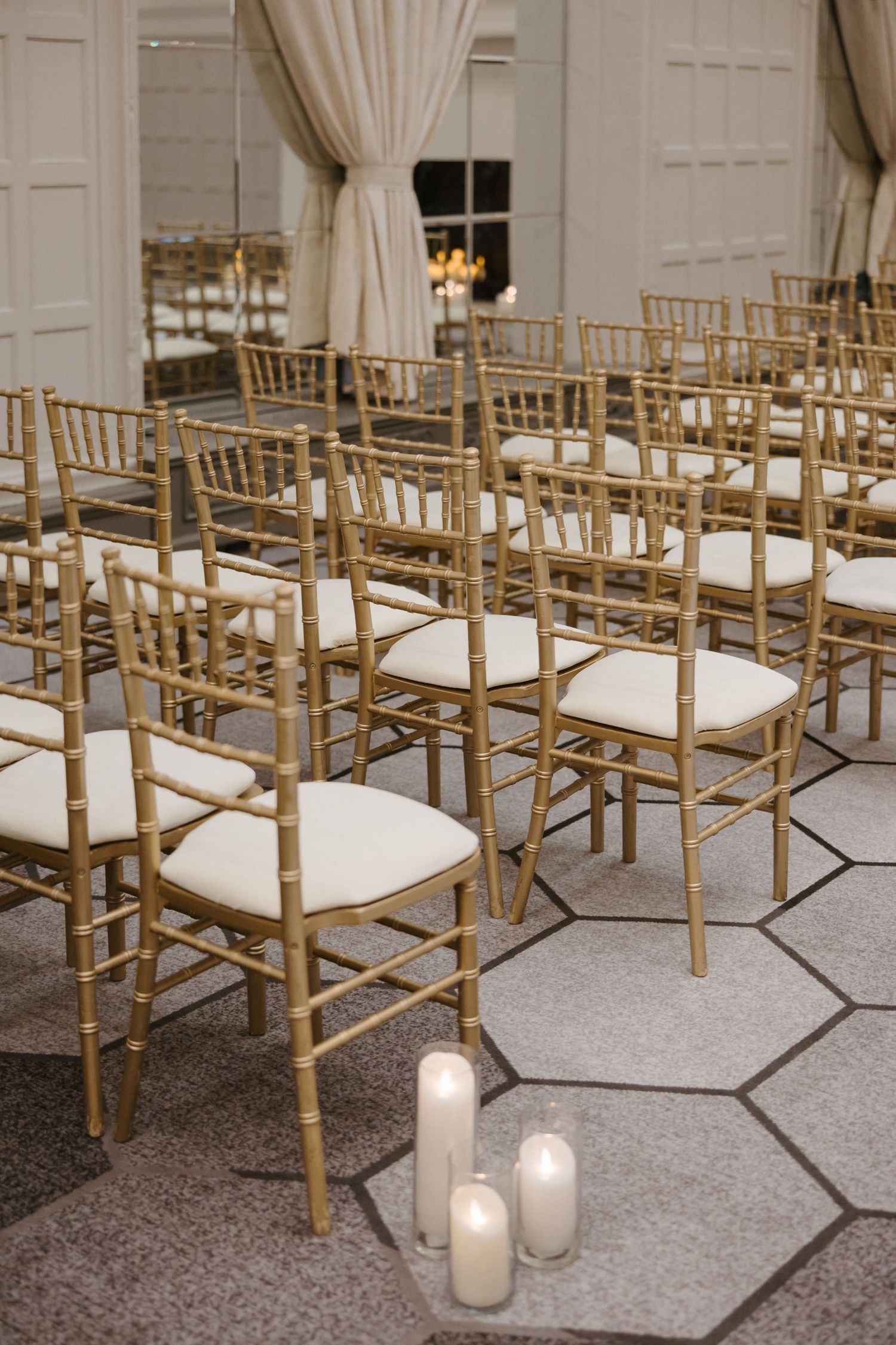 Gold chiavari chairs with white cushions arranged for a wedding ceremony, with pillar candles on hexagonal tile floor.