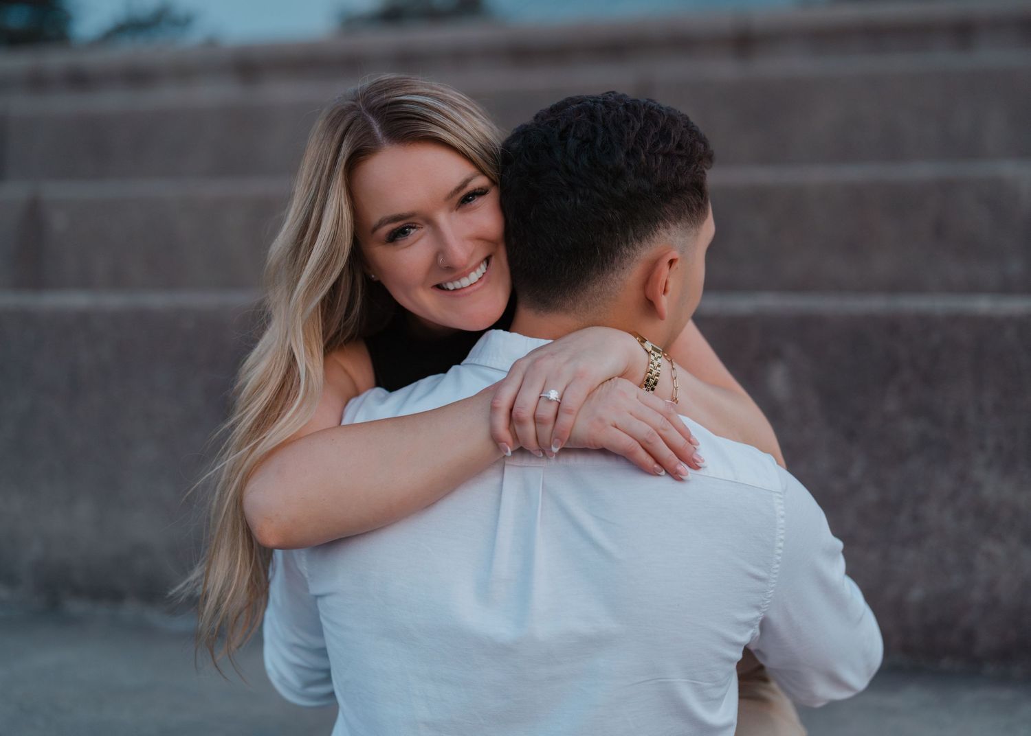 A smiling blonde woman hugs a man from behind on outdoor steps during an engagement photo session.
