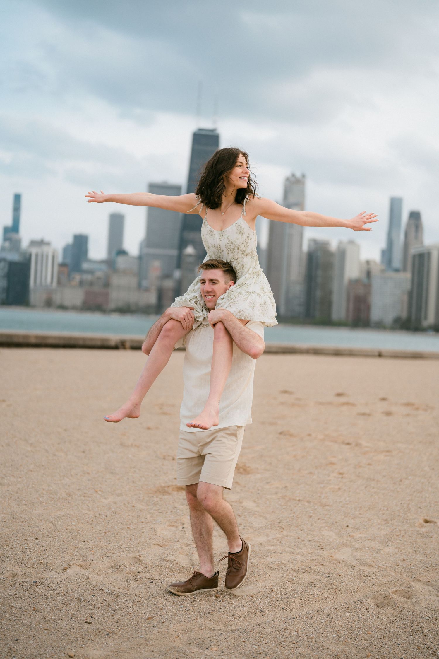 Man carrying woman on shoulders on Chicago beach, arms outstretched against cloudy sky.