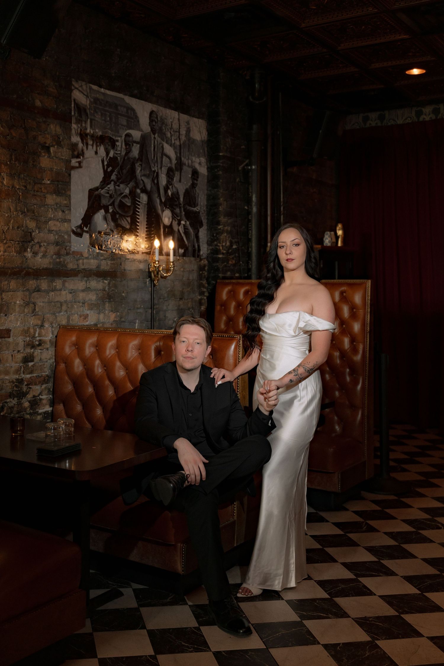 Couple poses in moody bar, man in black suit seated, woman in elegant white satin off-shoulder gown, checkered floor.