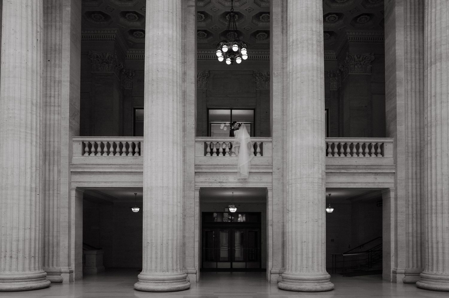 Black and white photo of wedding couple embracing on balcony between tall columns in grand neoclassical building.