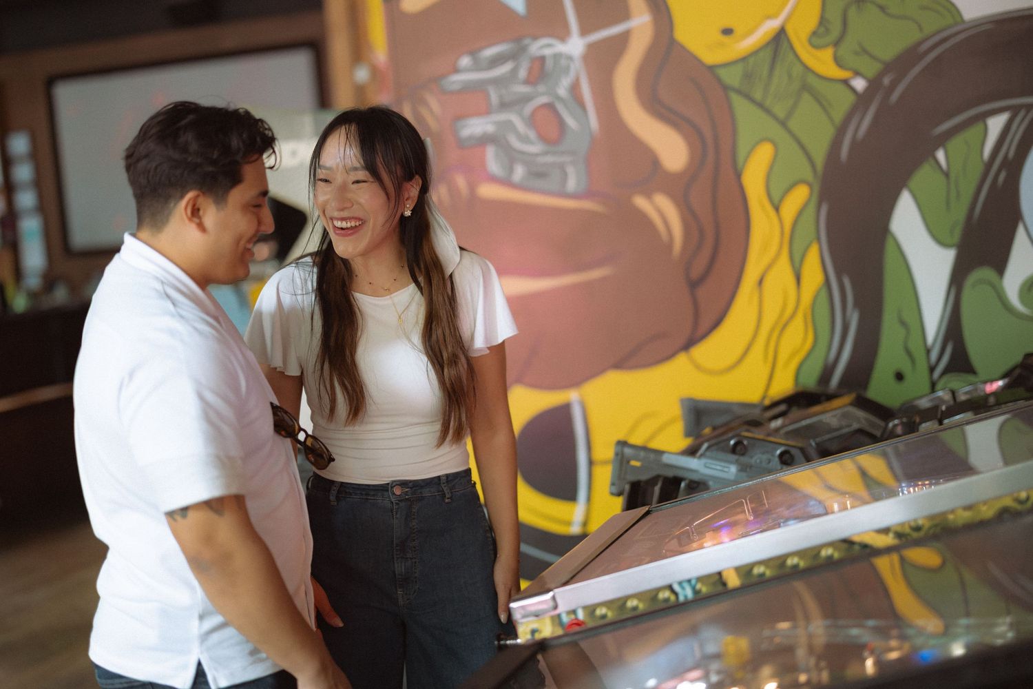 A smiling couple enjoys time together at an arcade, standing near a pinball machine with colorful wall art behind them.