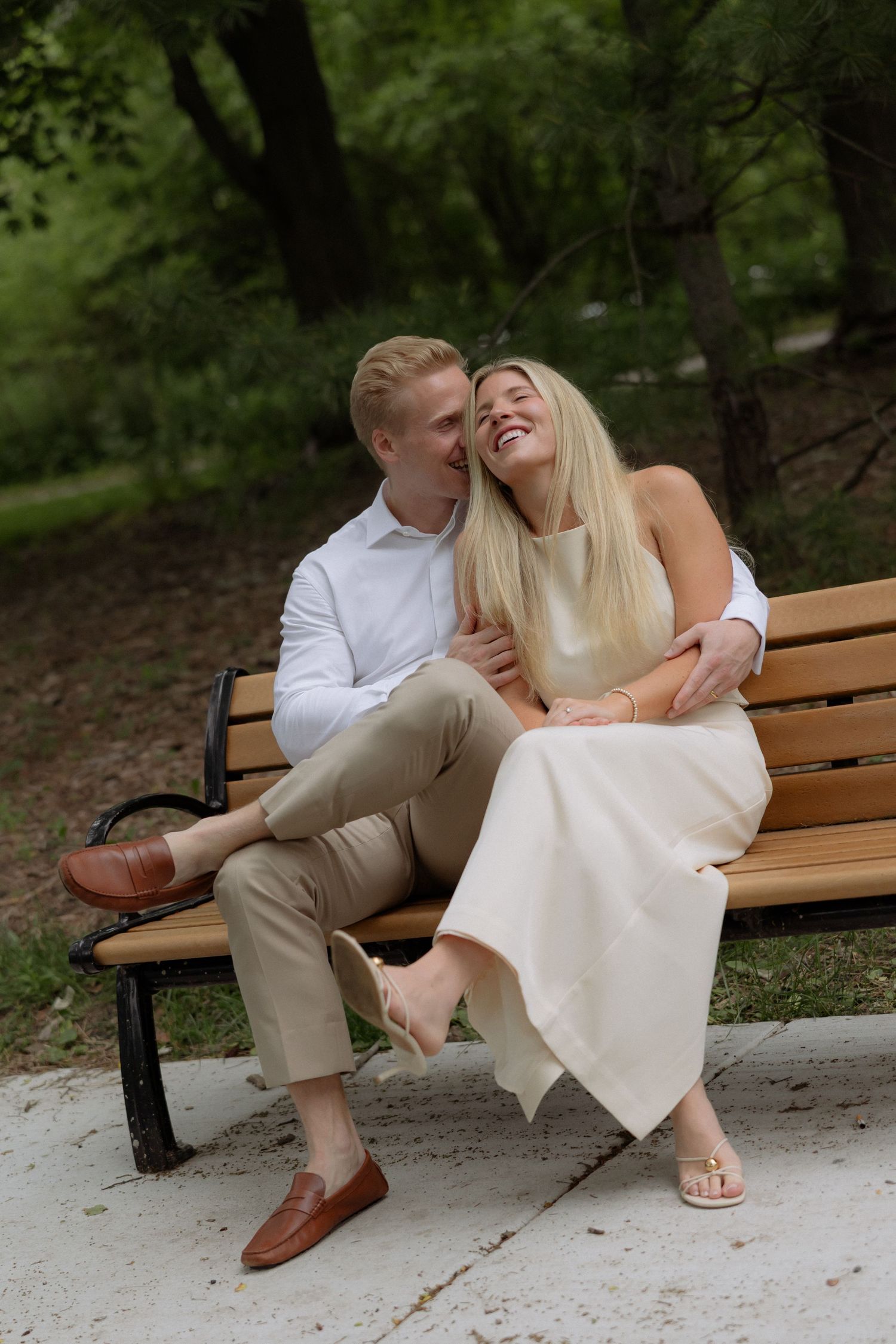Couple laughing together on a park bench, woman in cream dress, man in white shirt and khaki pants.