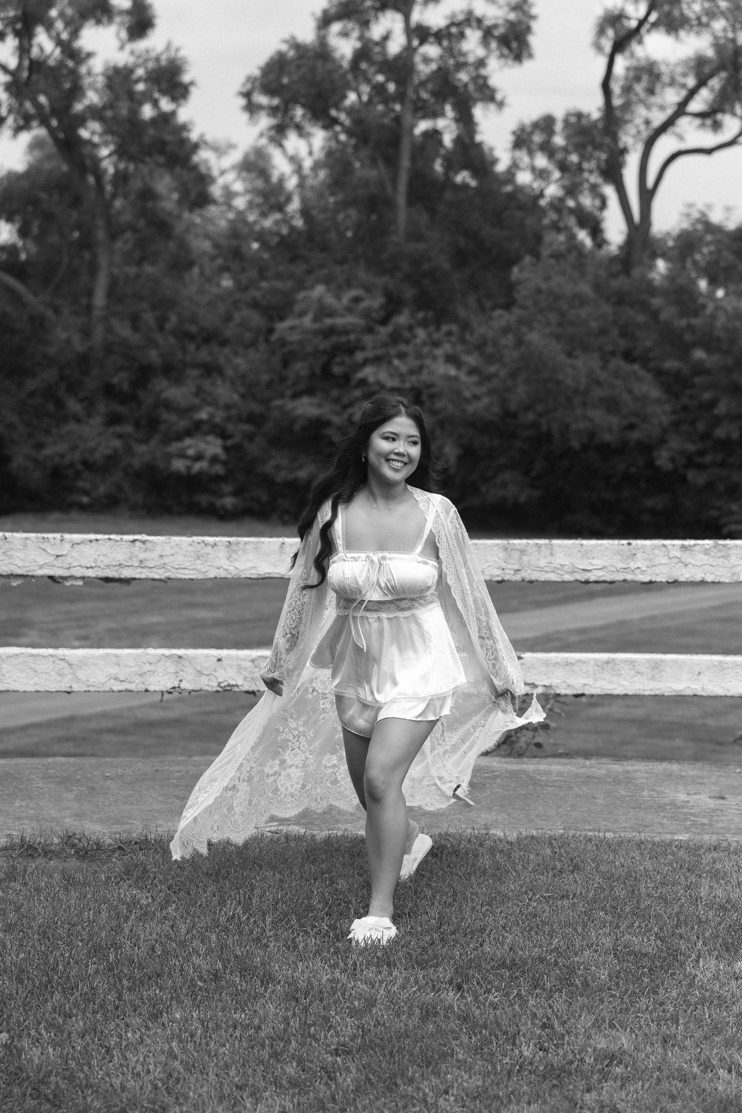 Young woman in white dress and lace kimono walks barefoot on green grass in black and white photo.