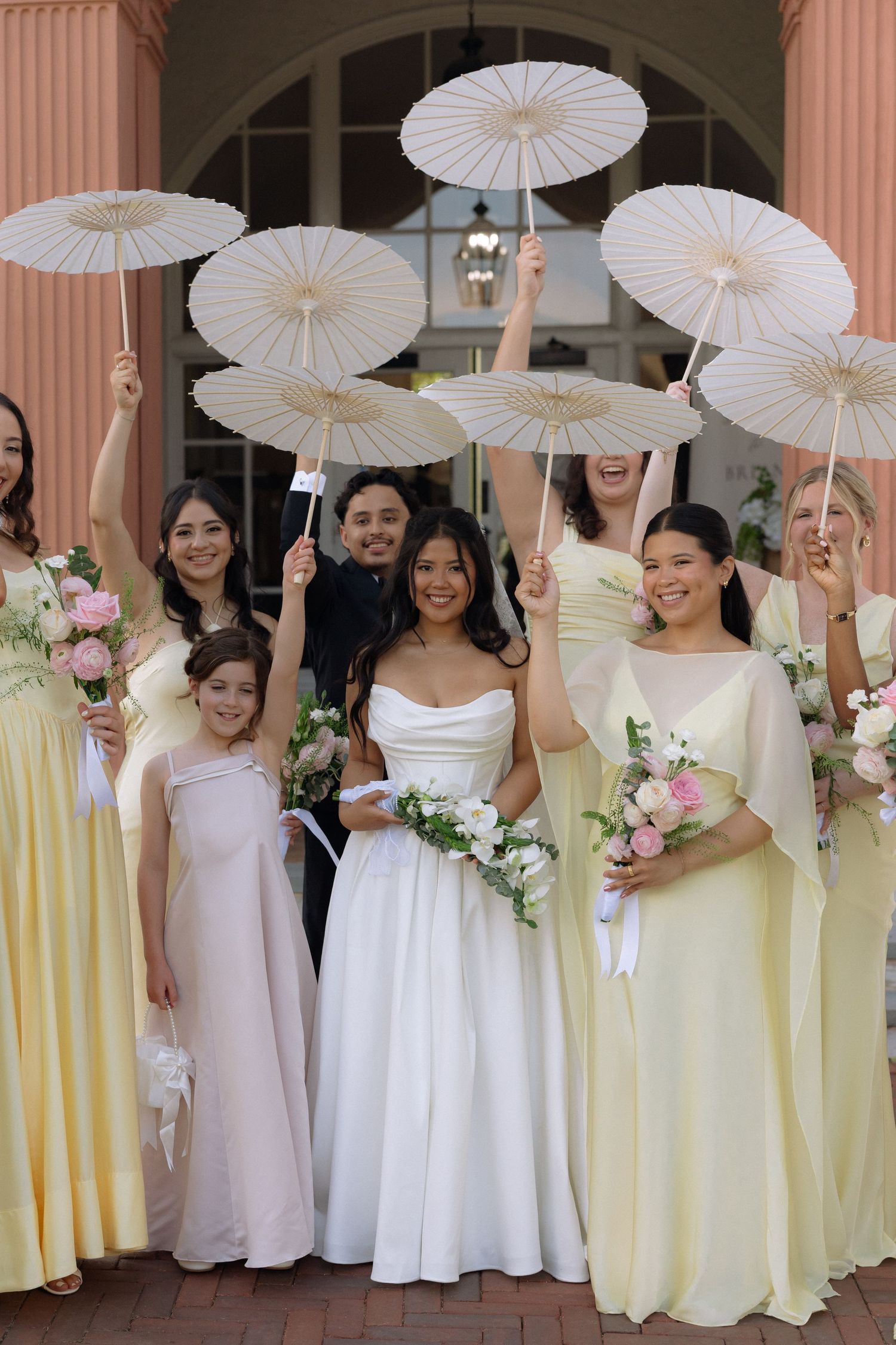 Smiling bride with bridesmaids in yellow dresses raising white parasols outside pink wedding venue.