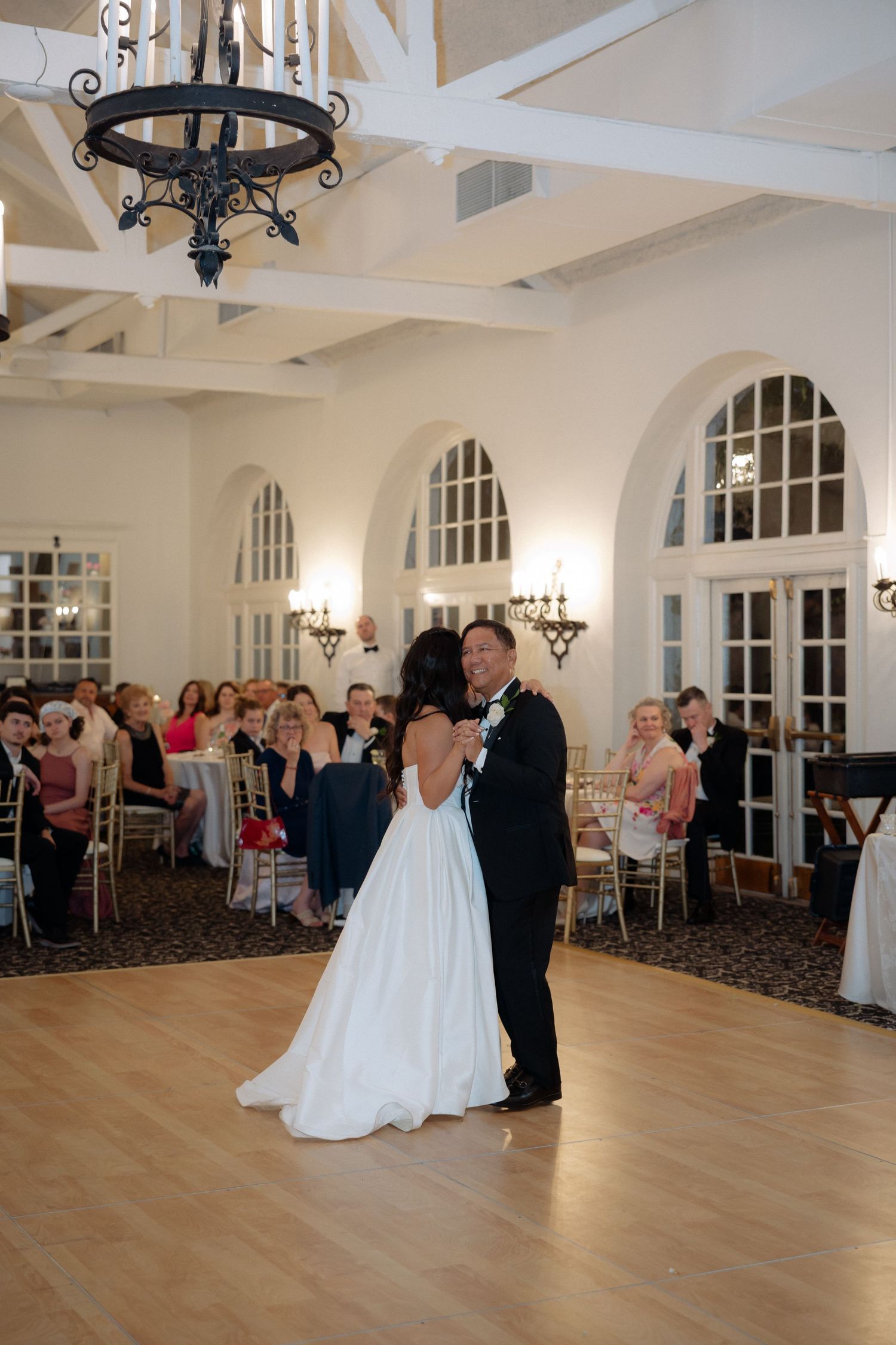 Bride and groom share their first dance in a beautiful white ballroom with arched windows and chandelier lighting.