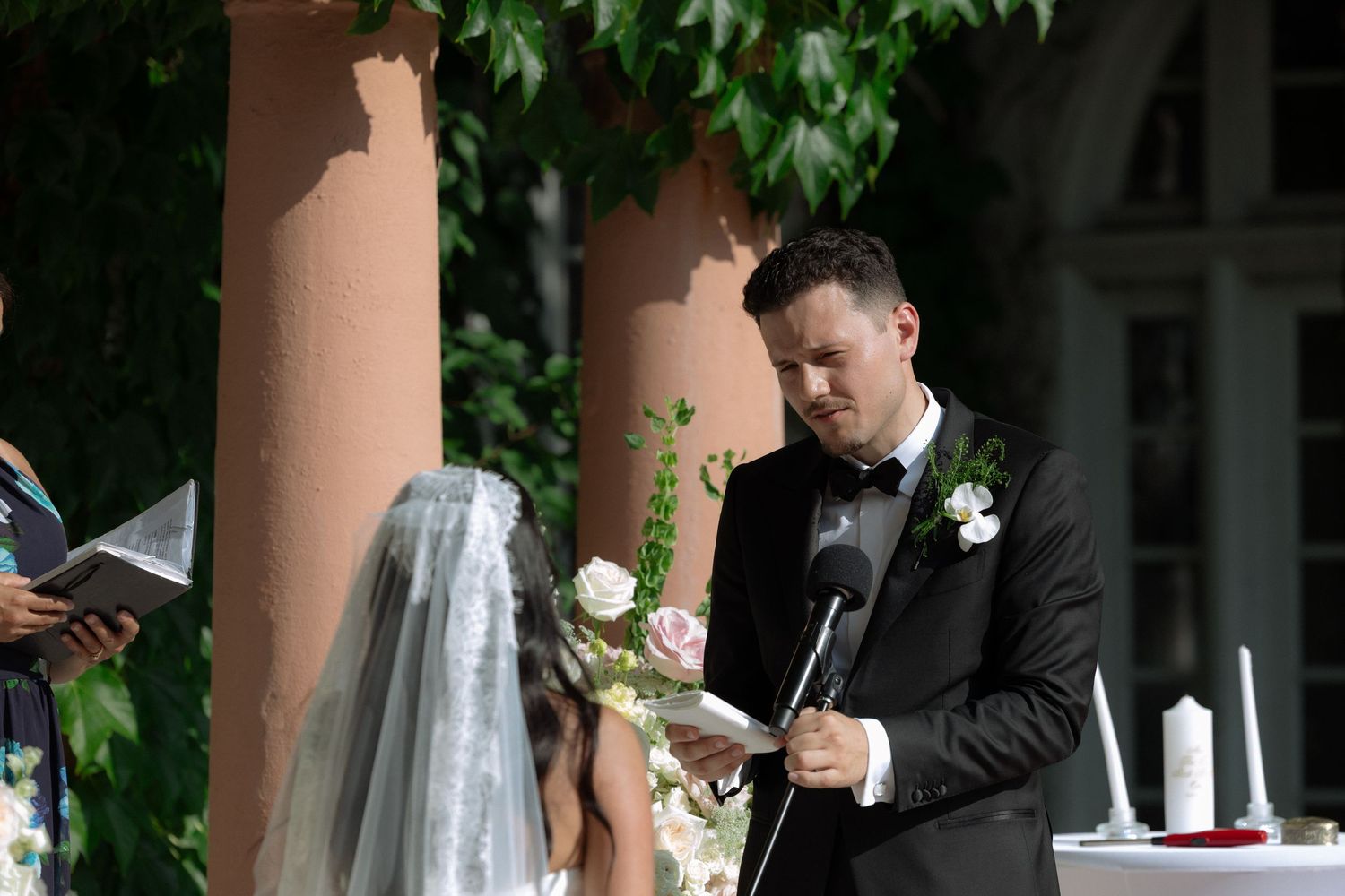 Groom in black tuxedo reads wedding vows outdoors at elegant ceremony with bride in veil and white roses.