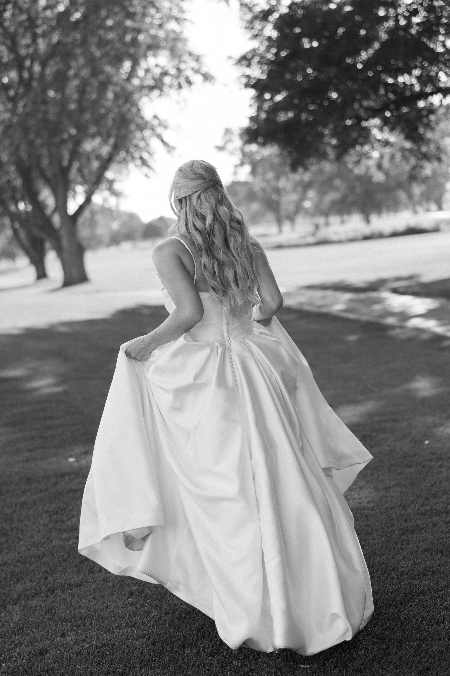 Black and white photo of a bride in a flowing ball gown walking through a lush green park, her long hair cascading down.