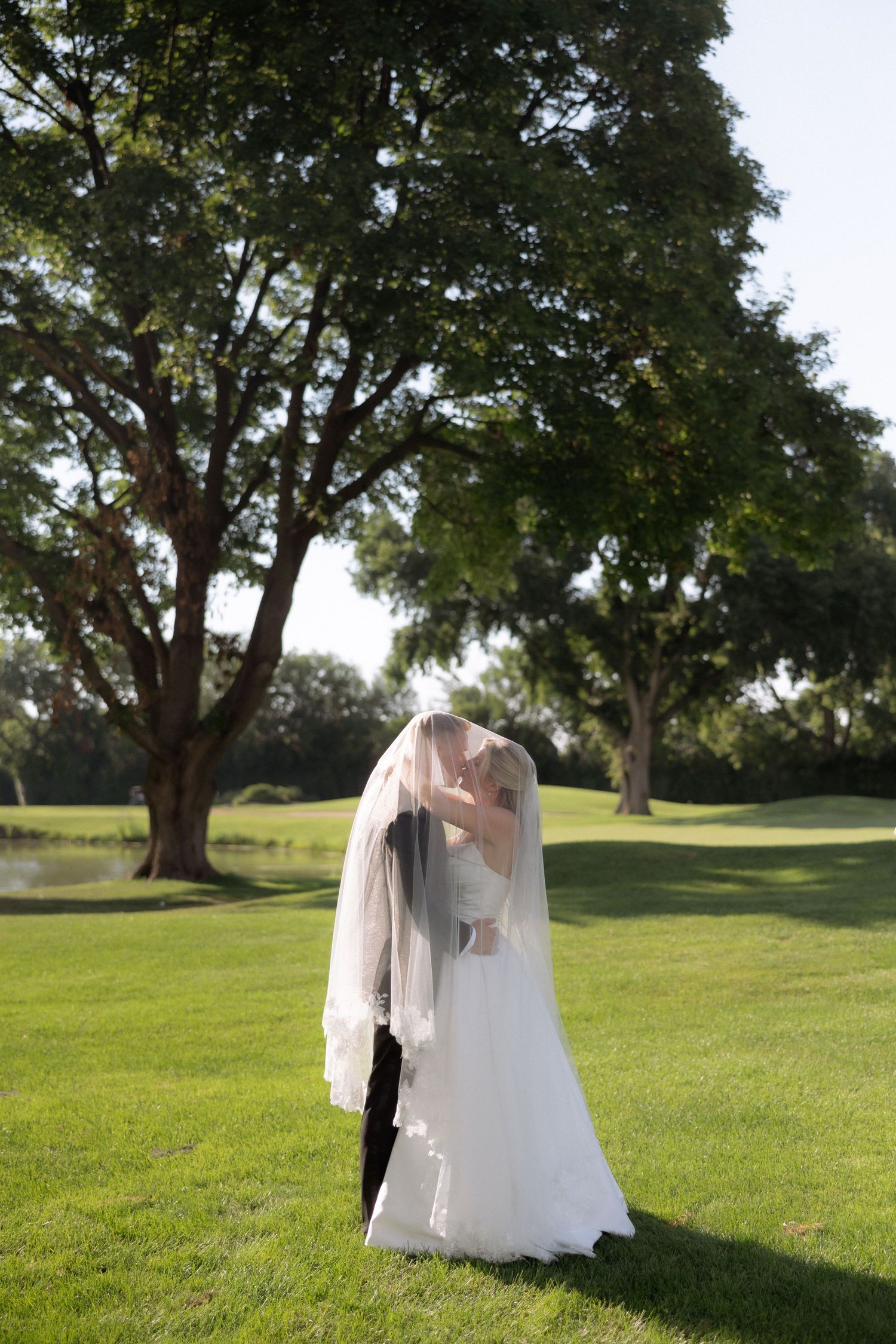 Bride and groom share romantic kiss under flowing veil on lush green lawn surrounded by tall trees.