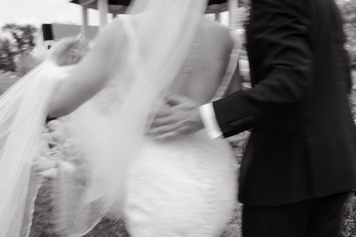 Black and white photo of a bride and groom walking together, her veil flowing in the wind on their wedding day.