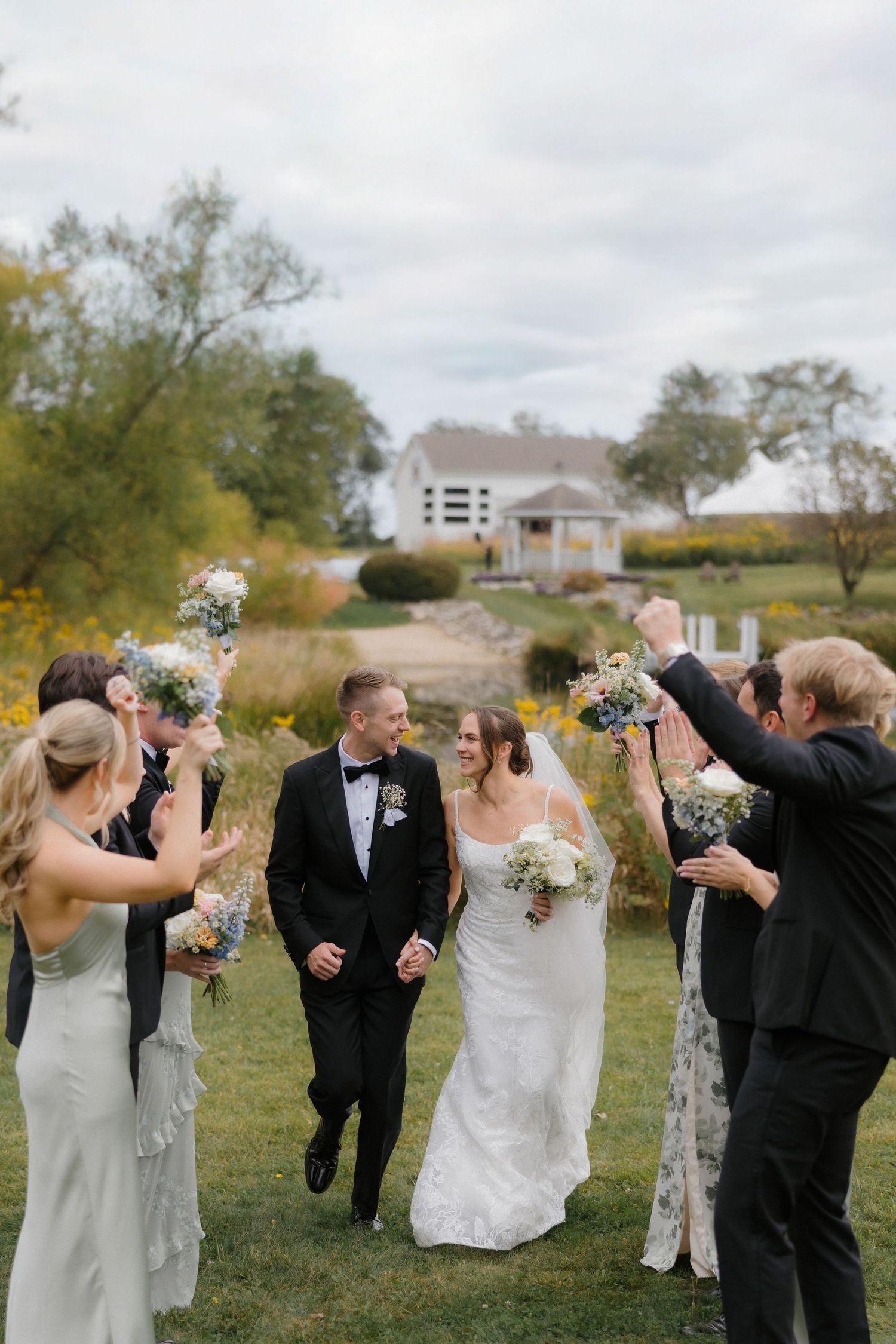 A bride and groom walk joyfully through a wedding party tunnel of raised bouquets on a lush green lawn.