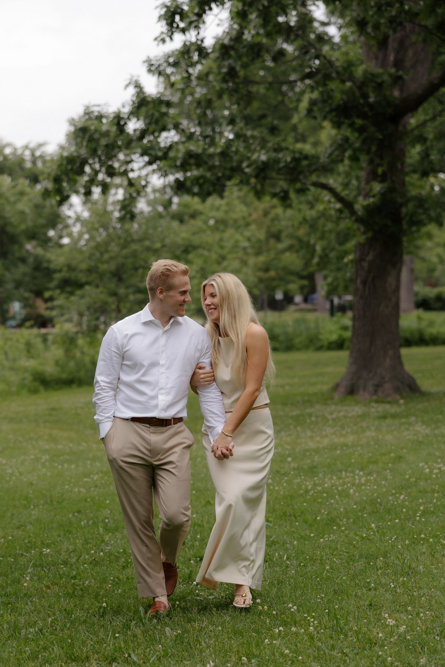 A smiling couple walks hand-in-hand through a lush green park, the woman in a satin dress and man in smart casual attire.
