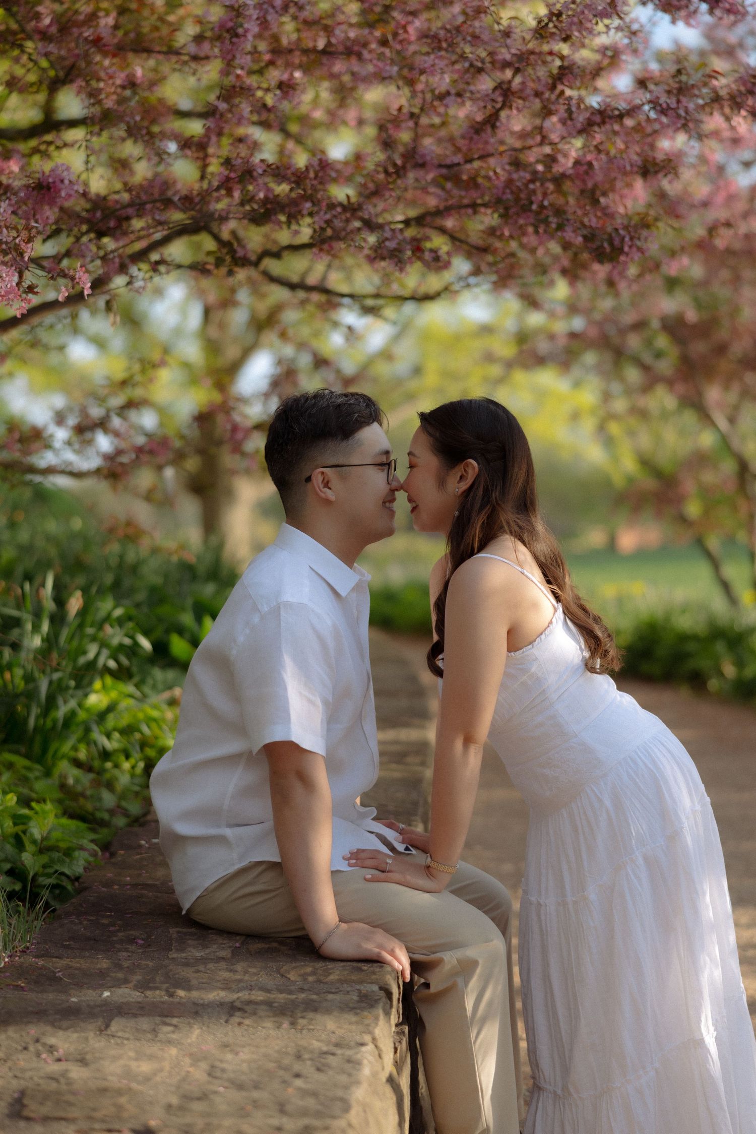 Couple sharing a romantic moment under blooming cherry blossom trees, woman in white dress, maternity photo session.