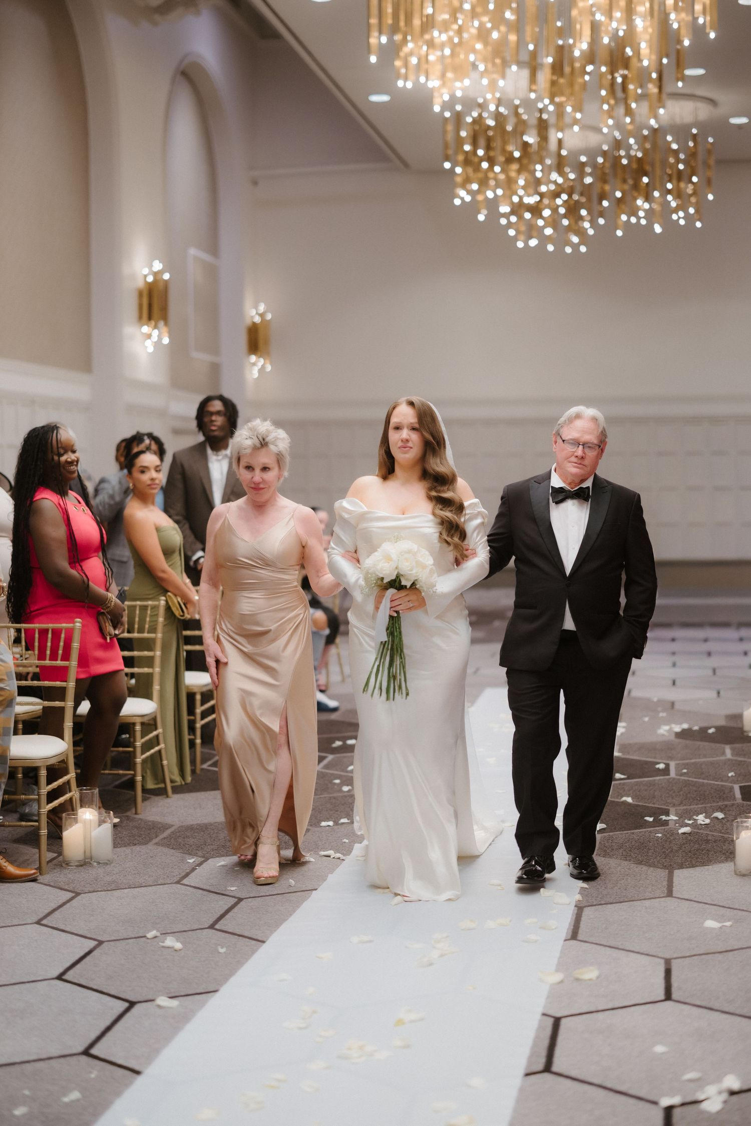 Bride walks down aisle with parents in elegant ballroom wedding ceremony, wearing white gown and holding bouquet.