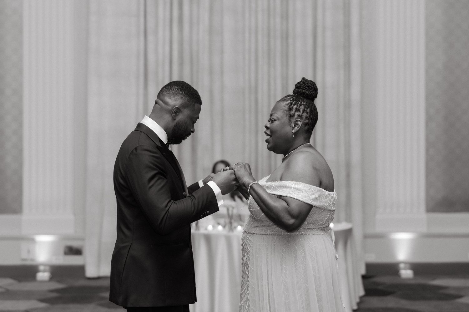 Black and white photo of a couple exchanging rings during a wedding ceremony in an elegant ballroom.