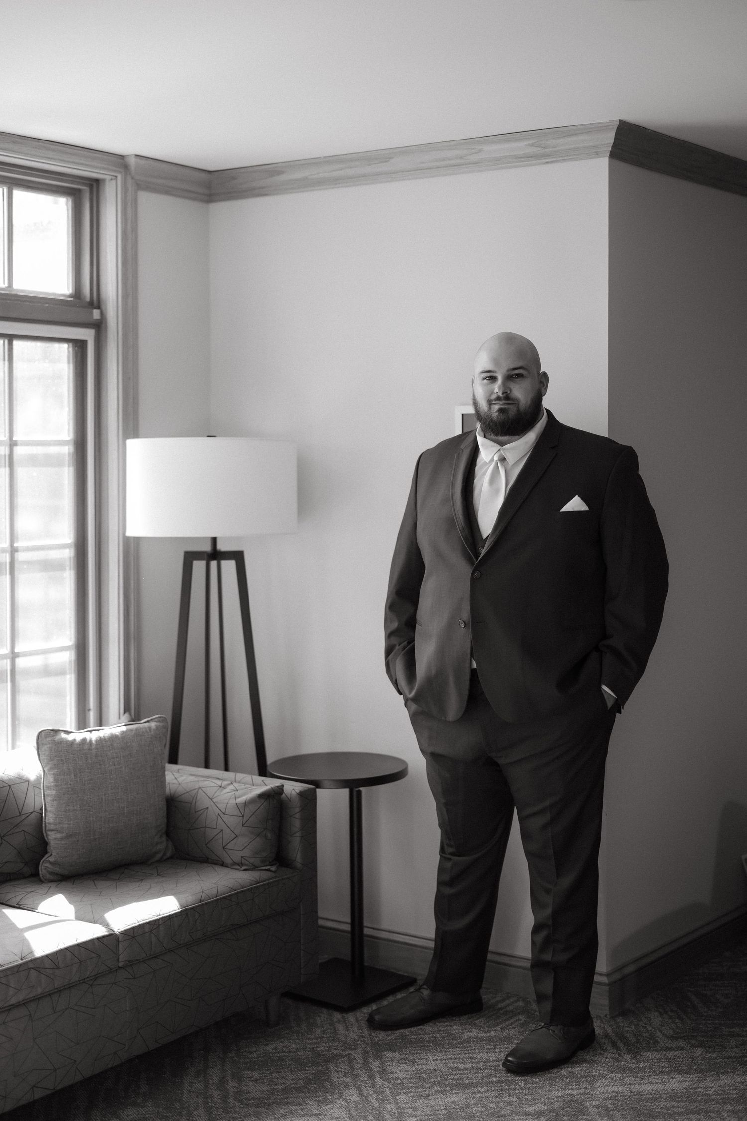 Bald bearded man in a dark suit with white pocket square stands confidently in a modern room, black and white photo.